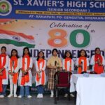 Students on stage at st Xaviers high school celebrating 80th independence day girls in white outfits with orange scarves indian flag backdrop